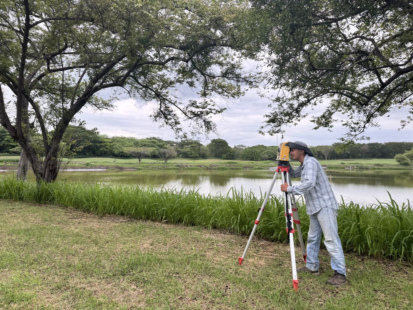 Surveyor measuring terrain beside a pond under guanacaste trees